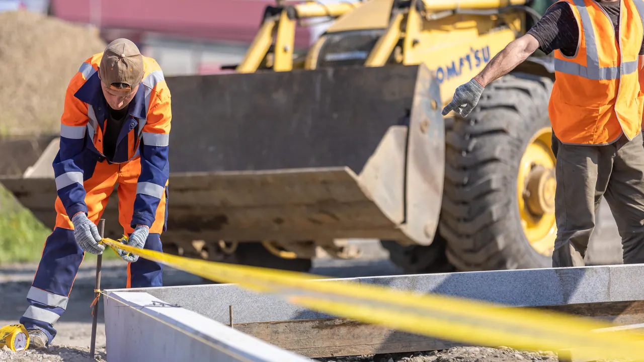 Dwaj pracownicy rozciągają miarę na placu budowy przy pracującej ładowarce