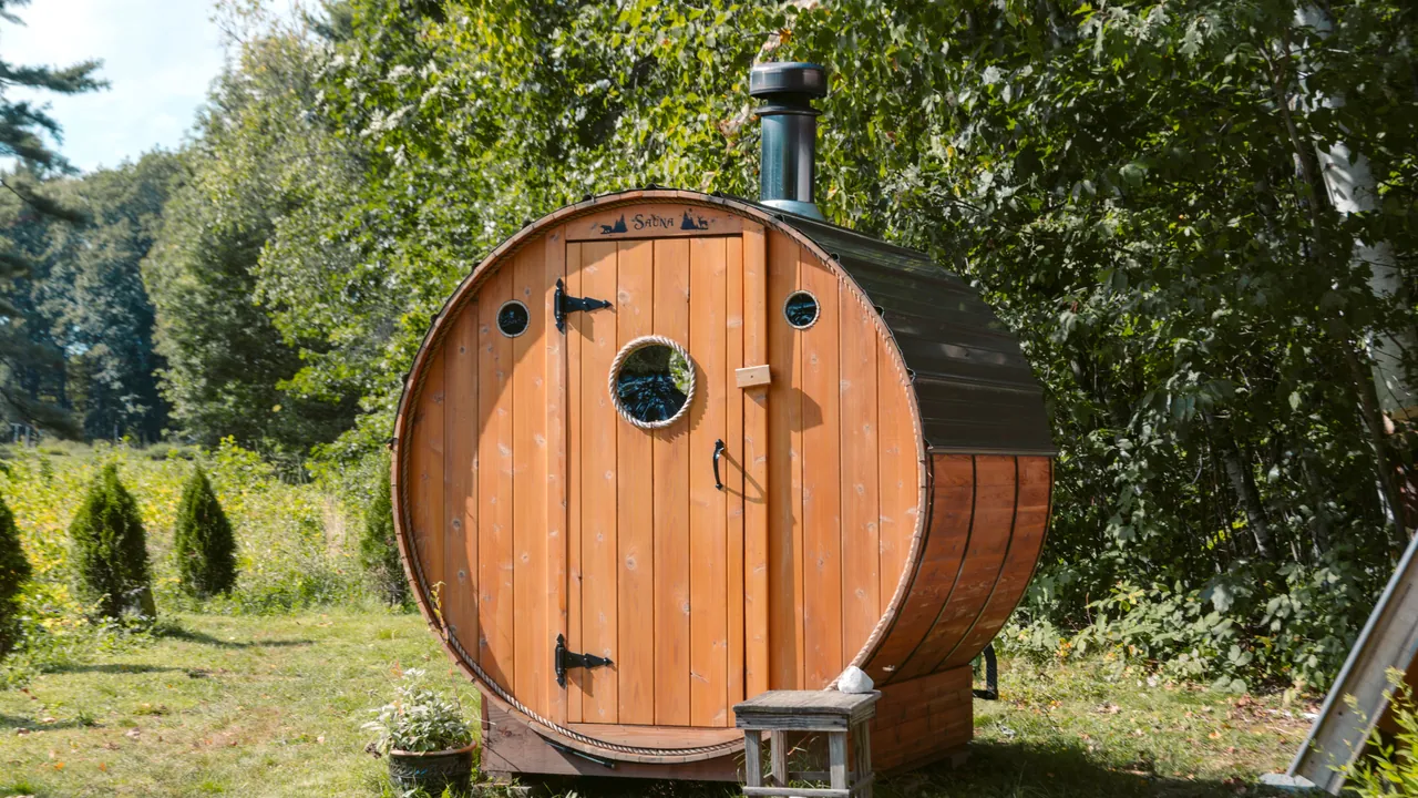Drewniana sauna w kształcie beczki z kominem, stojąca w ogrodzie przy tui